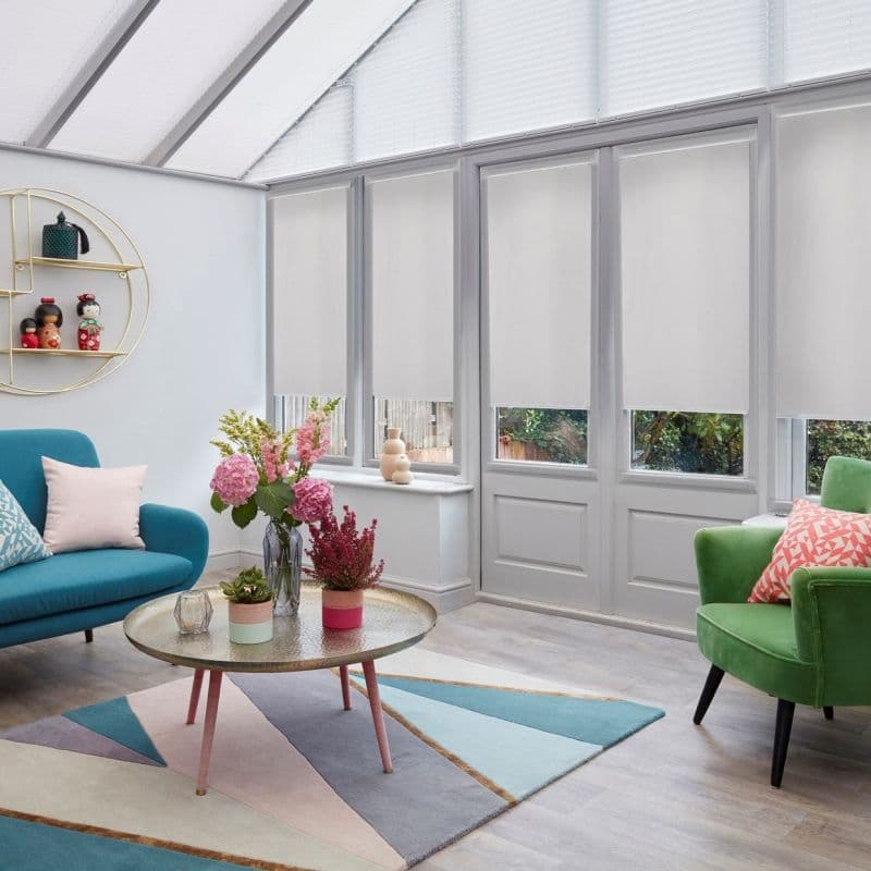 White roller blinds over tall glass doors and lower windows, lowered to mid-height filtering daylight, with pleated skylight shades, in a bright conservatory-style living room with colorful sofas, coffee table.