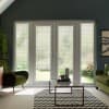 French double doors framed by sidelights admit diffused daylight through patterned blinds, centered in a living room with a glass coffee table on a chevron rug and olive-green armchairs.