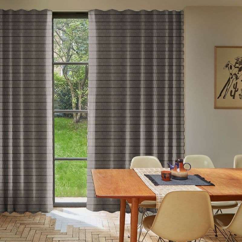 Floor-length patterned curtains with geometric print, drawn mostly closed leaving a narrow central gap showing a four-pane window; they filter soft daylight into a dining room facing a green garden.