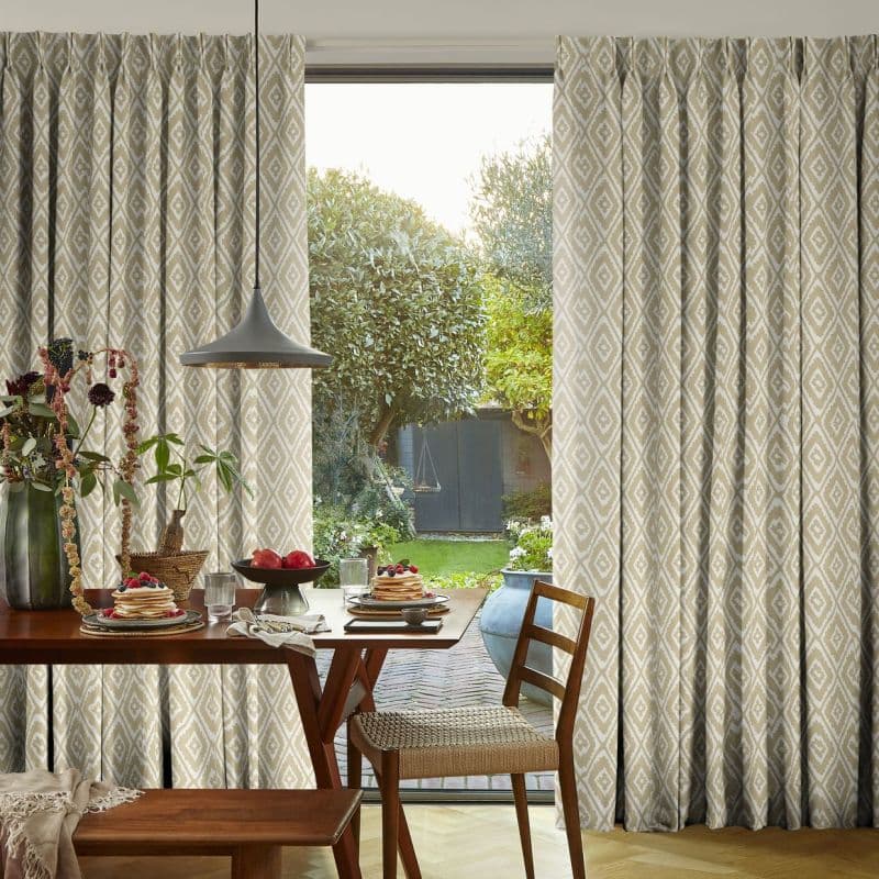 Beige diamond-patterned pleated curtains, full-length and parted at a sliding glass door, filtering soft daylight; wooden dining table set for breakfast in foreground, sunlit garden with trees and shed beyond.