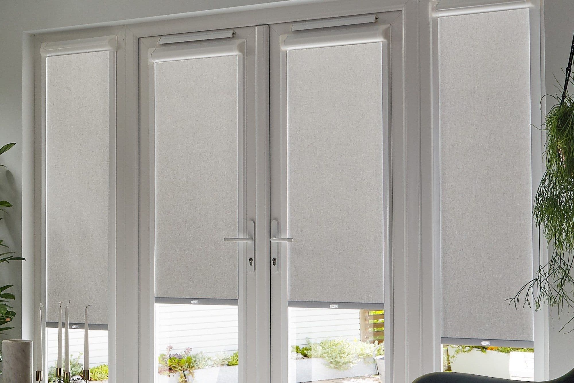 Grey roller blinds with top cassette housings across four tall window/door panels, lowered to filter light with narrow bottom gaps; central French doors, potted plants in a bright modern room.