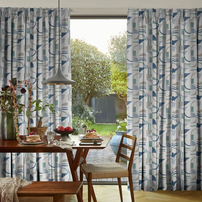 Floor-length pleated curtains with blue-and-teal geometric pattern frame a sliding glass door, pulled aside to reveal a sunlit backyard; wooden dining table with pancakes and pendant light in foreground.
