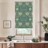 Teal-green floral roller blind hangs over a window above a stainless sink; light filters through. Wooden countertop holds a bowl of greens, potted plants, folded towel, and framed sketch.