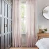 Sheer pale-pink, pleated full-length curtains, slightly parted at center, filtering soft daylight through a tall sash window; bedroom setting with dark paneled wardrobe, wooden bedside table, lamp, round mirror.