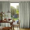 Full-length pale green textured pinch-pleat curtains, parted at center, framing and softening daylight; beyond a sunlit garden, foreground wooden dining table set with pancakes, vase, pendant lamp and chairs.