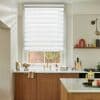 White dual-layer zebra roller blind lowered over window, alternating opaque and sheer horizontal bands filtering daylight; above a brass kitchen faucet, marble countertop and wooden cabinets with brick-and-plant courtyard visible.