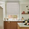 A zebra roller blind, lowered across a window with alternating opaque and sheer stripes, filtering daylight over a kitchen sink with brass faucet, wooden cabinets and marble countertop.