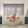Grey horizontal Venetian blinds covering a large dormer window, slats mostly closed and filtering soft daylight. In front: wooden desk, pink swivel chair, lamp, plant in a bright attic study.