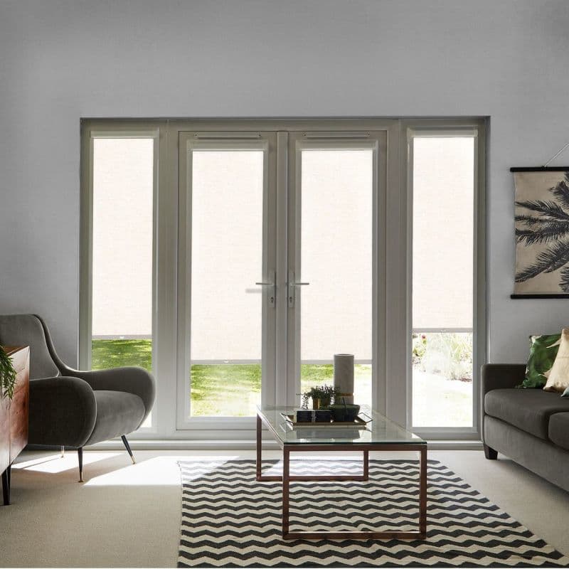 Four white roller blinds covering French doors and sidelights, lowered and diffusing daylight; fronting bright living room with mid-century armchair, sofa, glass coffee table on chevron rug, glimpse of lawn.