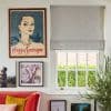 Grey fabric Roman blind (layered fold, pull chain at right) half-lowered, filtering daylight. Context: bright living room with framed art, red sofa, potted plant; outside hedge and brick. Text: Maggy Sarragne