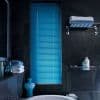 Blue horizontal Venetian blinds, closed, their slats letting narrow light strips through a narrow window in a dark slate-tiled bathroom with bathtub, folded towels, toiletries and a turquoise vase.