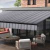 A diagonal-striped retractable fabric awning with a striped valance is fully extended from the house eave, shading a glass-walled patio furnished with low table, sofa and armchairs in daylight.