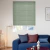 Sage-green pleated cellular blind, lowered with thin horizontal ribs and bottom edge slightly above the sill, blocking light; in a sunlit living room with a blue sofa, cushions, and a floor lamp.