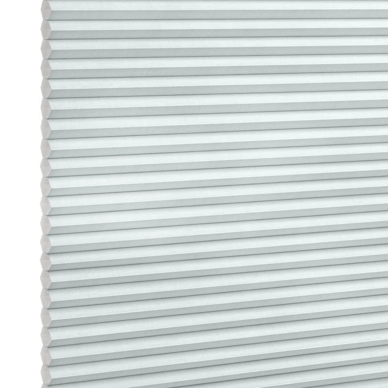 Pleated honeycomb blind, pale gray-blue with hexagonal cells along the left edge, fully lowered and closed, softly filtering daylight, set against a bright white background in a neutral-lit interior.