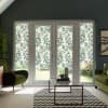 French patio doors covered with translucent leafy-patterned film, filtering daylight into a living room with green velvet chairs, glass coffee table, zigzag rug, and potted plants.