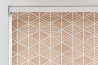 A fabric roller blind with white geometric diamond lattice over peach woven texture, mounted in a white cassette and lowered (closed), filtering light in a bright, neutral interior window recess.
