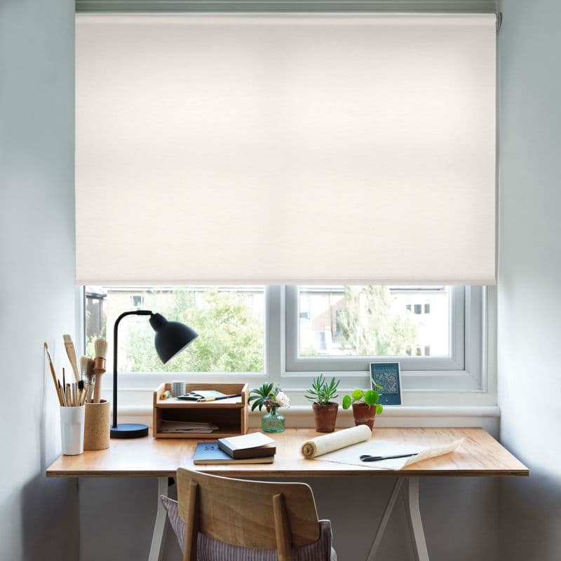 White fabric roller blind, fully lowered across a double window, softly diffusing daylight; small wooden desk beneath holds a lamp, brushes, notebooks and potted succulents in a light, calm home workspace.