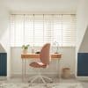 White horizontal Venetian blinds across a wide window, slats tilted with fabric tapes; filtering bright daylight and casting striped shadows; attic home-office niche with wooden desk, pink swivel chair, lamp.