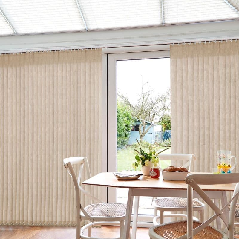 Beige vertical blinds covering patio doors, slightly parted at center and softly filtering daylight; small dining table with wicker‑seat chairs, vase of flowers and bread, glimpse of backyard.
