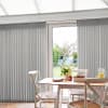 Vertical fabric blinds, light gray, partly parted to reveal a central glass patio door, slats hanging straight; filtering soft daylight into a dining nook with wooden table, white chairs, garden view.