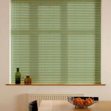 Pale green horizontal blinds, lowered and closed, diffusing daylight and obscuring the view; below them a sill with two bottles, a radiator, and a table with a bowl of oranges.
