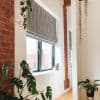 Gray Roman blind, partially lowered with soft folds, covering a black-framed window and filtering daylight. Bright minimalist living area with exposed red brick columns, wooden floor and potted plants.