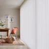 White vertical blinds covering a floor-to-ceiling window, closed and softly filtering sunlight; dining nook with wooden table, chair with terracotta cushion, patterned rug, wicker basket, botanical prints, bright daylight.