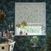 A leaf-printed Roman blind, mostly lowered across a white-framed window, filtering daylight; surrounded by dark palm wallpaper, houseplants, and a small bar cart.