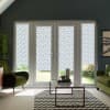 Glass double doors with patterned blinds blocking and diffusing daylight, in a modern living room with green armchairs, glass coffee table, chevron rug, and a view of the garden.