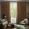 Patterned rust-orange, floor-length pleated curtains framing a sash window, pulled slightly apart to reveal bright greenery; they filter soft daylight into a cozy living room with plush sofa, pouf, chair.