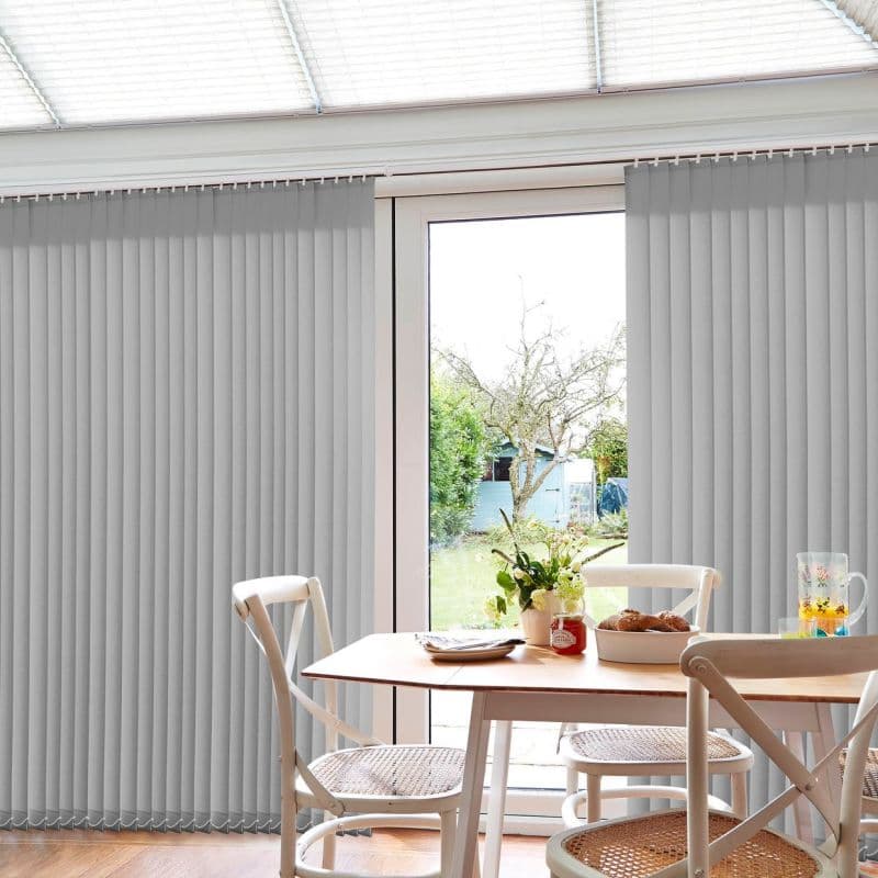 Light-gray vertical blinds, full-height, partially parted at a central glass door, filtering daylight; a dining table with white chairs, vase of flowers and pastries in a room overlooking a garden.