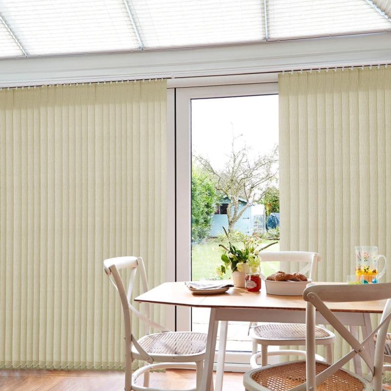 Beige vertical blinds, floor-length with a central opening; mostly closed, filtering soft daylight while parted at the glass door; bright dining nook with table, white chairs, vase, pastries, garden beyond.