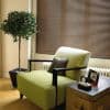 Brown venetian blinds cover the window, closed and softly filtering daylight; in a cozy living-room corner with a green armchair, trunk side table, potted topiary and hardwood floor.