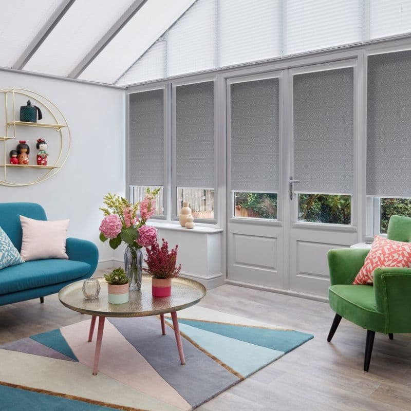 Grey patterned roller blinds on four tall French-door windows, lowered to mid-height and filtering light while leaving a narrow lower view; bright conservatory living room with pastel furniture and flowers.