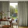 Full-length patterned curtains, beige with green fern print, parted to reveal sliding door; filtering soft daylight into a dining nook with wooden table set for breakfast and garden views.