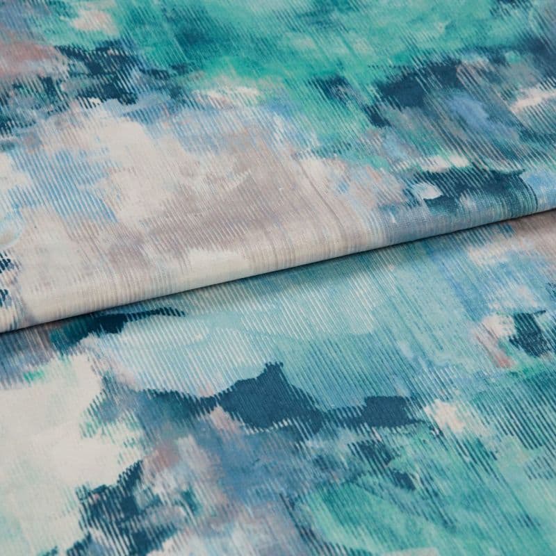 A folded ribbed fabric panel with watercolor-like teal, blue, and grey splotches; lying overlapped and slightly rolled, close-up on a flat surface with soft, diffuse lighting.