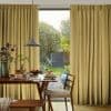 Floor-length pleated beige curtains, slightly parted at a sliding door, filtering daylight; wooden dining table set with pancakes and fruit beneath a pendant lamp, opening onto a lush backyard.