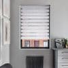 White zebra roller blind, lowered with alternating opaque and sheer horizontal bands; filtering light and obscuring view; in a gray kitchen nook with espresso machine, radiator below, brick building outside.
