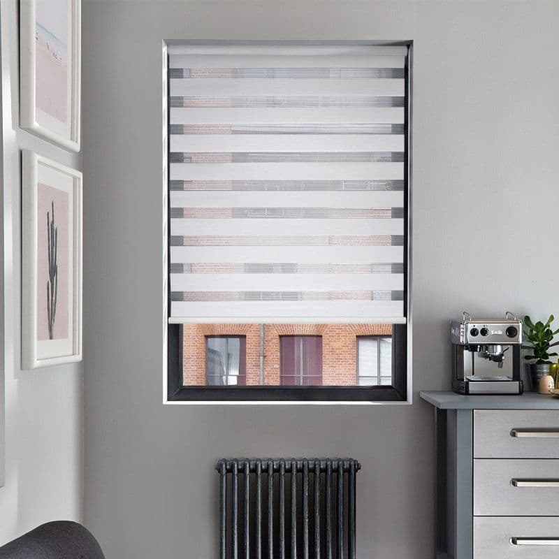 White zebra roller blind, lowered with alternating opaque and sheer horizontal bands; filtering light and obscuring view; in a gray kitchen nook with espresso machine, radiator below, brick building outside.