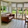 Light-grey roller blinds, lowered halfway across two windows and a sliding glass door, gently filtering daylight; overlooking patio with table and chairs, leather sofa in bright living-dining room.