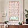 Floral-patterned roller blind hangs down over a kitchen window, filtering daylight; stainless-steel faucet and sink below, countertop with bowl of greens, potted plants, and a framed sketch nearby.