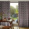 Patterned pleated curtains with geometric maroon and grey blocks, pulled aside, framing an open glass patio door and filtering daylight; dining table with breakfast setting and garden view.