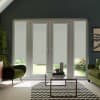 Glass patio doors with four white roller blinds lowered, filtering daylight into living room with green armchairs, glass coffee table on black-and-white chevron rug and potted plants.