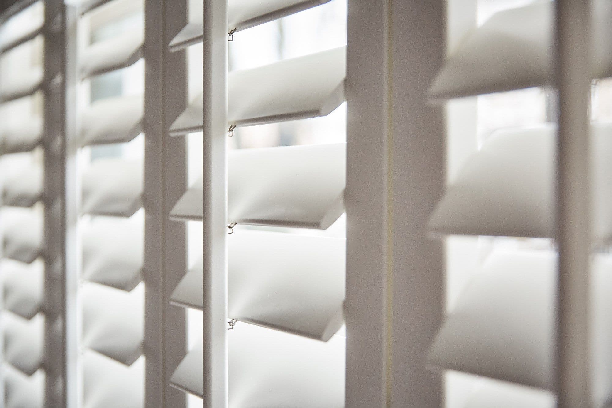 White plantation-style louvered shutters, close-up showing vertical stiles and thick horizontal slats, are partially open, angling to softly filter daylight into a bright, blurred interior.