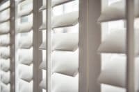 White plantation-style louvered shutters, close-up showing vertical stiles and thick horizontal slats, are partially open, angling to softly filter daylight into a bright, blurred interior.