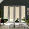 Beige roller blinds cover three-pane French doors and sidelights, blocking sunlight while revealing a narrow garden strip below, in a dark-walled living room with green armchairs and glass coffee table.