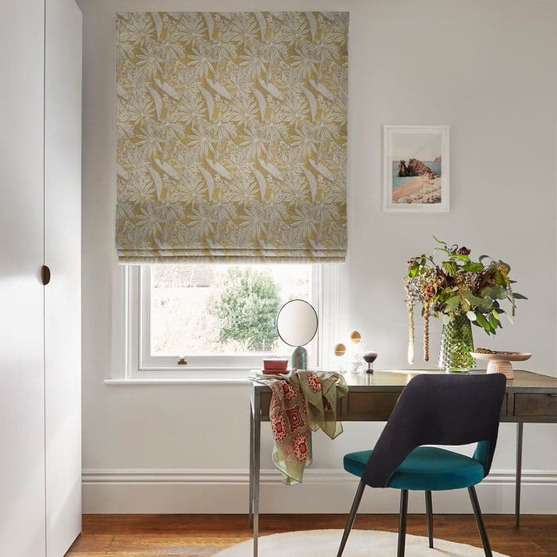 A patterned yellow-and-white Roman blind, lowered into soft folds, filtering daylight through the window; above a wooden desk with teal chair, vase of flowers, scarf and mirror in a bright room.