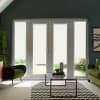 Double glass patio doors with lowered white roller blinds diffuse daylight into a living room, centered by a glass coffee table on a chevron rug between two green chairs, garden beyond.