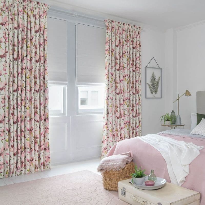 Floral pleated curtains, drawn to the sides over closed white Roman blinds, filtering soft daylight into a pastel bedroom with a pink bedcover, wicker basket and botanical wall art.