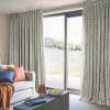 Patterned, floor-to-ceiling pleated curtains, drawn partly open at a sliding glass door, filtering bright daylight into a cozy living room with sofa, cushions, coffee table and a fenced backyard view.
